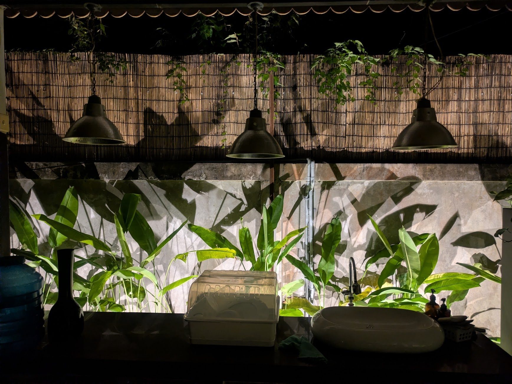 An outdoor kitchen at night: three pendant lamps over wall-washed plants
